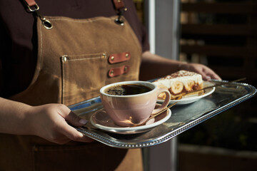 Caucasian young adult woman holding silver tray serving cup of black coffee and slice of cake, only torso and hands visible, wearing apron, standing indoors near window