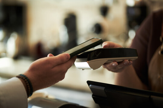 Caucasian young adult man holding smartphone making contactless payment using digital wallet at point of sale terminal, female hand operating card reader in retail environment