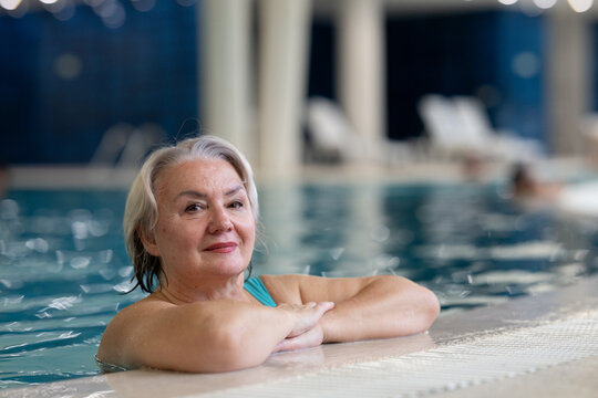 Smiling senior woman enjoying relaxation in an indoor swimming pool, leaning on the poolside. Concept of active lifestyle, wellness, health, hydrotherapy, and leisure for older adults.