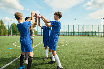 Group of teenage boys and one teenage girl with diverse ethnicities celebrating together on outdoor soccer field, raising hands for high five after successful play during practice