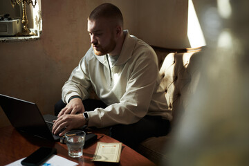 Fototapeta premium Caucasian young adult man sitting at table using laptop computer, focusing on typing with hands on keyboard, glass of water and notebook nearby, natural indoor setting