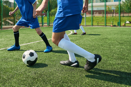 Teenage boys playing soccer on outdoor field, focusing on legs and feet in motion while competing for ball, athletic activity taking place during sunny day, green artificial turf visible