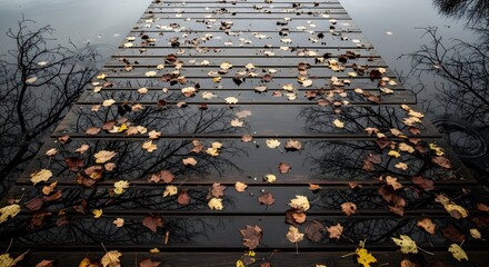 Melancholic autumn scene with wet wooden pier and fallen colorful leaves reflecting bare tree silhouettes in the dark water, creating a moody seasonal background