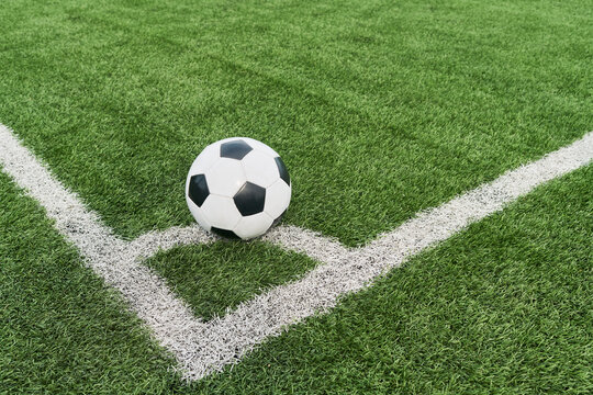 Soccer ball resting on corner of green field near boundary lines, positioned for corner kick, showing close up of sports equipment on outdoor artificial turf