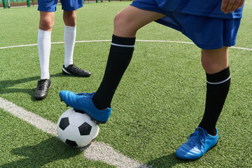 Two teenage boys standing on soccer field, one teenager resting foot on soccer ball while other teenager standing nearby, both wearing sports uniforms and soccer cleats