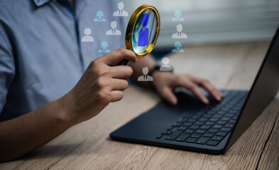 Fototapeta premium Hand holding magnifying glass in front of laptop with digital human icons. Concept of HR recruitment, talent search, employee hiring, business workforce, and candidate selection