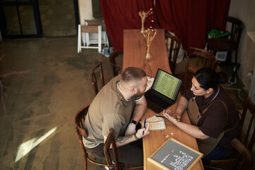 Caucasian young adult man and Hispanic young adult woman sitting at table discussing menu options with laptop and menu board, woman wearing apron, man holding pen and notepad