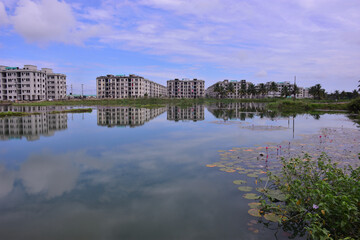 Fototapeta premium Modern residential apartment buildings reflected in a calm pond, with blooming wildflowers and greenery in the foreground. A scenic blend of urban development and natural beauty in Bangladesh