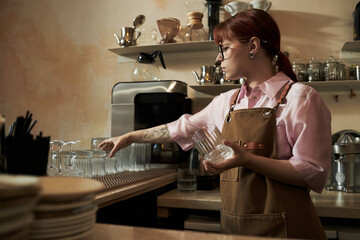 Young adult Caucasian woman with red hair and tattoos arranging clean glasses behind counter in cafe, wearing apron and eyeglasses, focusing on organizing glassware in workspace