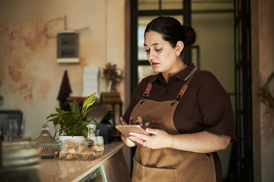 Young adult Hispanic woman standing behind counter using digital tablet, focusing on screen while managing orders in cafe or bakery, wearing apron, hair tied back