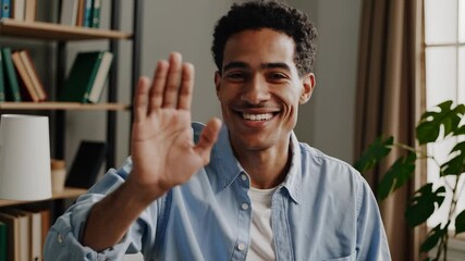 The man wears a blue shirt while the man looks to the side in a room. The man is seated near bookshelves and greenery while the man remains indoors by the window. - Powered by Adobe