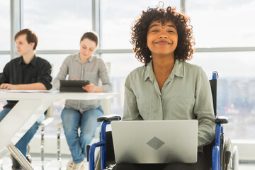 Accessible environment. African woman in chair for people with disability working with laptop in modern office. Diverse multiracial group of people colleagues working together. Young professional team