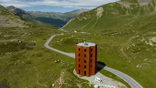Aerial view of the rusty red tower standing firm amidst the winding road and rolling green hills, a contrast to the distant, stony mountains, Julier Pass, Grisons, Switzerland.