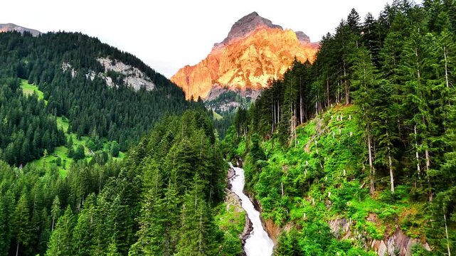 A scenic view of Lenk, Simmenf&auml;lle in Switzerland with a waterfall flowing through dense green forest, tall pines on steep slopes, and a towering mountain glowing in the warm light of sunset