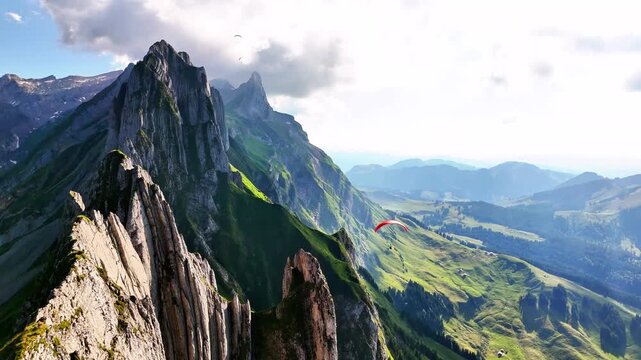 Aerial view of Sch&auml;fler Switzerland featuring jagged alpine ridges, lush green valleys, and paragliders soaring above dramatic mountain peaks under partly cloudy skies