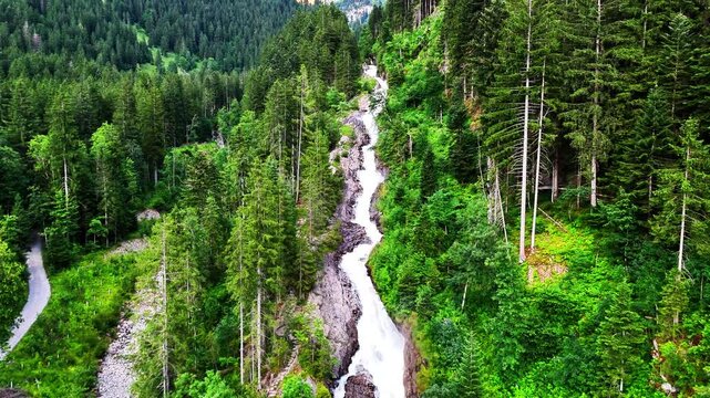 Scenic alpine waterfall flowing through dense green forest in Lenk, Simmenf&auml;lle, Switzerland, surrounded by tall trees and mountain peaks glowing in warm sunlight against a clear sky