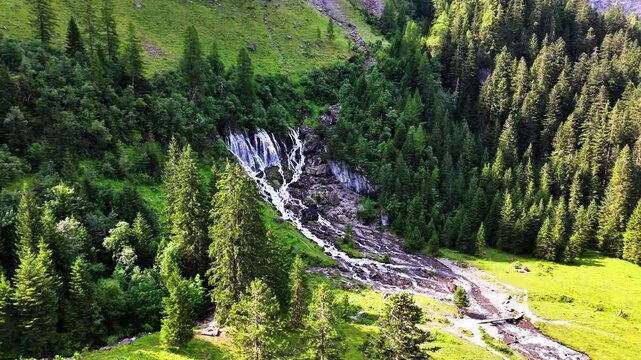 Aerial view of Lenk Sibenf&auml;lle in Switzerland, featuring a cascading alpine waterfall flowing through rocky terrain surrounded by lush green forest, tall pine trees, and vibrant mountain meadows