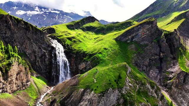 The Engstligenf&auml;lle waterfall in Adelboden, Switzerland, plunges from steep alpine cliffs into a vibrant green valley, surrounded by meadows, rugged mountains, and traces of snow in the distance
