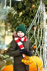 Obraz premium Happy cute teenager boy with mesh bag of tangerines standing near by spruce Christmas tree. Holiday celebrate concept.