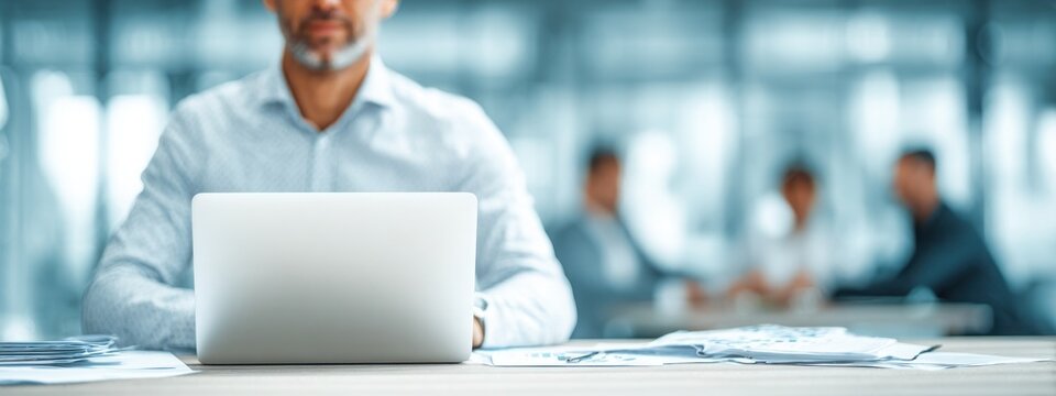 Close-up of businessman at desk with charts and laptop, blurred colleagues in background, representing corporate teamwork and data analysis.