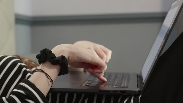 Female hand wearing scrunchie typing on laptop keyboard while resting comfortably on bed with striped sheets, representing modern remote work and digital communication lifestyle