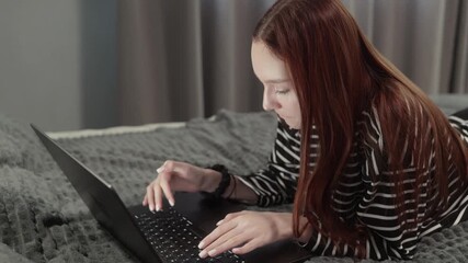 Redhead teenage girl comfortably lying on bed, typing on laptop keyboard while relaxing in casual home environment, engaging with digital technology and online communication - Powered by Adobe