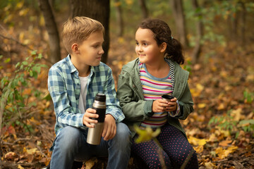 Kid teenager boy and girl hiking in the forest relax sitting on stump and drinks from thermos in autumn forest