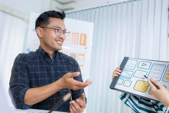 Three people are sitting at a table with a whiteboard behind them. One of the people is holding a pen and looking at a diagram on the board. Scene is focused and serious
