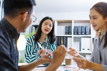 Three people are sitting at a table with a whiteboard behind them. One of the people is holding a pen and looking at a diagram on the board. Scene is focused and serious
