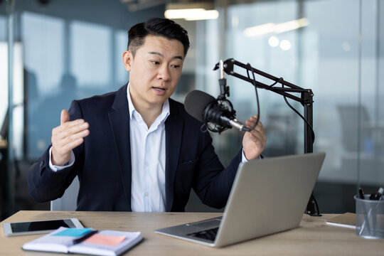 Young Asian man in a suit sits at an office desk and speaks into a microphone, holding an online meeting on a laptop