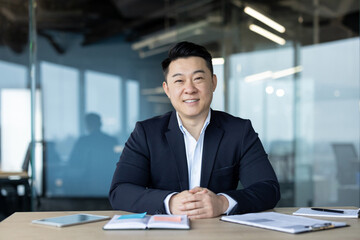Portrait of a young smiling and successful Asian businessman in a suit sitting at a desk in the office and looking at the camera