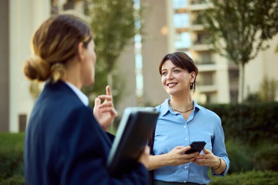Two Professional Women Chatting Outdoors in City Park, Smiling While Sharing Phone and Documents