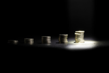 Front view of coins stacked at different heights with a spotlight highlighting the tallest coin,...
