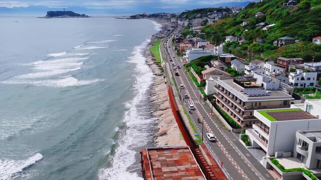 Aerial view of Kamakuro town in Japan showing a coastal road, ocean waves, a hillside residential area, and an island connected by a bridge.