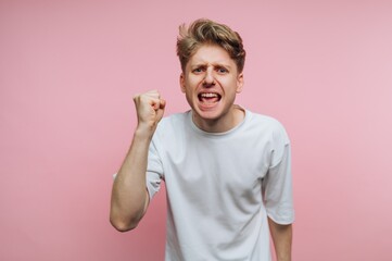 Fototapeta premium A young man in a white t-shirt expressing anger with a fist raised against a pink background.
