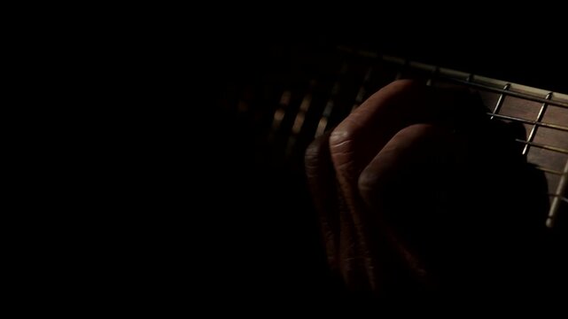 Cinematic close-up of a musician's hand playing chords on an acoustic guitar in a dark room with dramatic, moody low-key lighting.