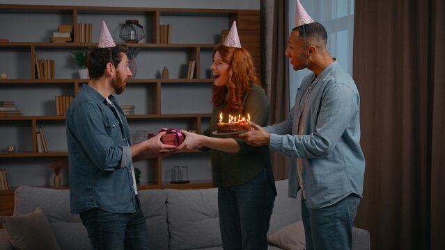 Three multiracial friends at home party birthday congratulation couple Caucasian woman with African American male guy congratulate man giving present box gift cake with candles celebrate together