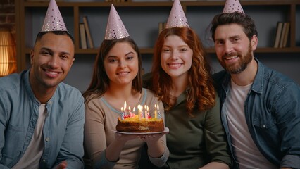 Four multiracial friends in party caps celebrate birthday holding cake smiling looking at camera portrait festive event at home diverse buddies carefree faces together posing indoors people men women