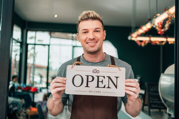 A smiling man in an apron stands in the doorway of his cafe, holding up a wooden "OPEN" sign. The sign also has Thai text and the word "WELCOME."