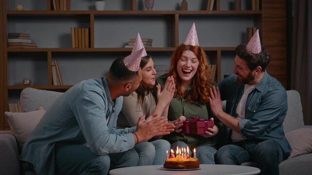 Four multiracial friends celebrating birthday party presenting present Caucasian woman receive gift from diverse friendly buddies men women cuddling applauding happy smiling congratulate event at home