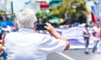 Older man captures a city street parade using his phone, viewed from behind on a bright, sunny day.