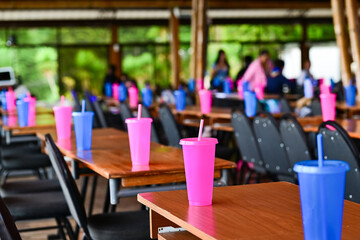Pink and blue plastic cups with straws are neatly placed on wooden tables in a spacious event hall or gathering space.