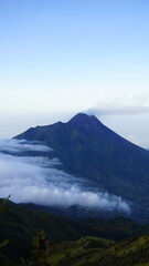 A majestic volcanic mountain peak rises above a thick blanket of white clouds under a clear blue sky.