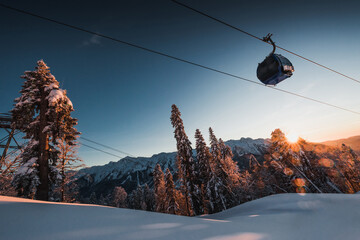 A gondola and ski lift against the backdrop of a winter landscape of mountains, trees, and an evening sunset in Krasnaya Polyana.