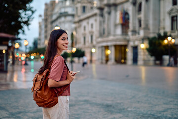 Young woman with backpack and smartphone smiling while exploring an urban street at dusk, enjoying travel and evening sightseeing between buildings