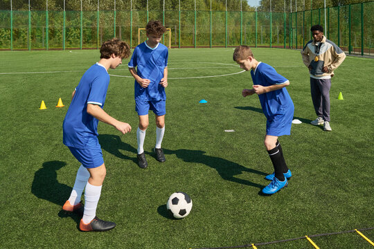 Three teenage boys practicing soccer dribbling drills on outdoor field while Black male coach observing training session, green fence and trees in background