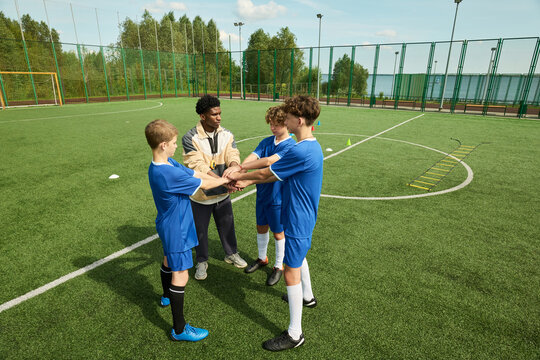 Black man coaching group of boys on soccer field, teenage boys standing in circle stacking hands together, coach leading team building exercise during outdoor practice