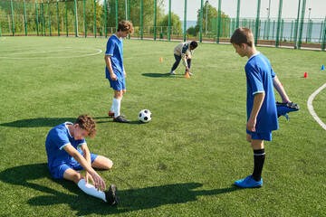 Group of teenage boys playing soccer on outdoor field, one teenager sitting on grass stretching leg...