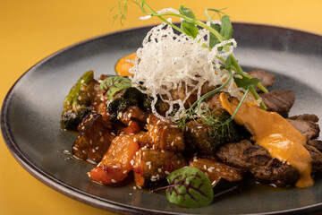 Colorful gourmet dish featuring grilled vegetables, tender beef slices, and a delicate garnish, beautifully arranged on a dark plate against a vibrant yellow background