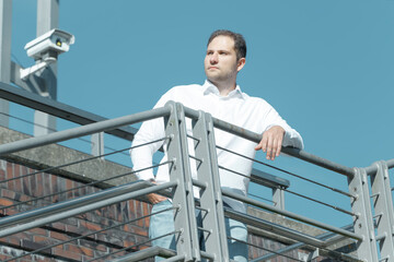 A young businessman in a white shirt and blue jeans stands on a street staircase with metal railings. Summer. Blue sky. Caucasian.